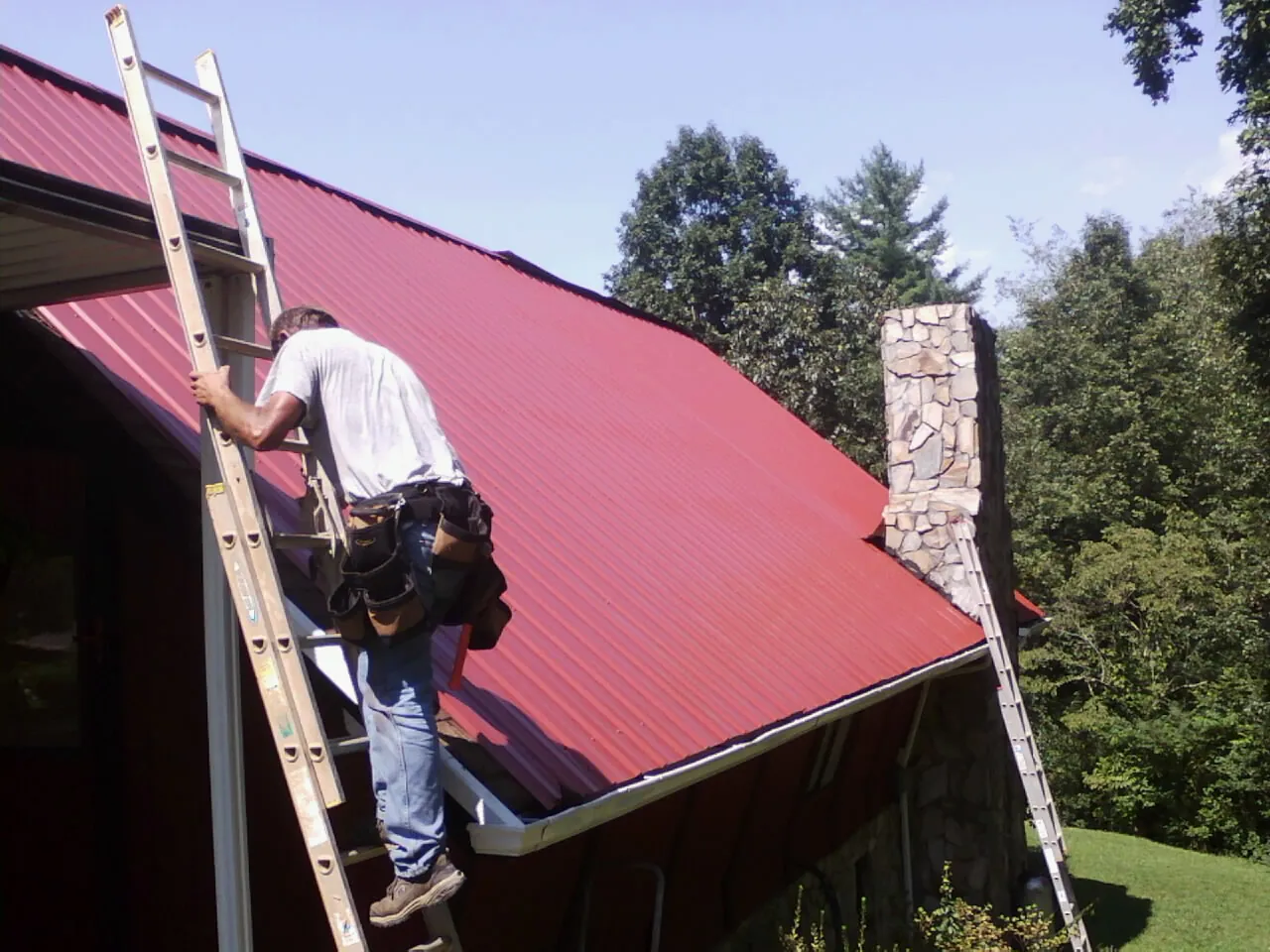 Roofer on a ladder beside a red metal roof and stone chimney — Soffit & Fascia Repair by Roofing Brothers in Trenton